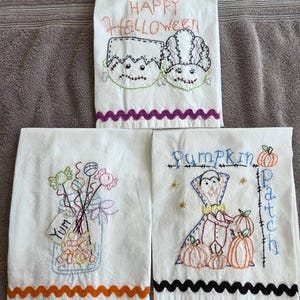 May include: Three white Halloween-themed kitchen towels with embroidered designs. One towel says "Happy Halloween" with monster faces. Another has a jar of candy, and the third says "Pumpkin Patch" with a vampire and pumpkins. Each towel has a decorative border.