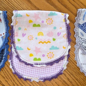 May include: Three baby bibs with different patterns. One has robots, another has stars and rainbows, and the third has zebras and giraffes. Each bib has a patterned bottom and is trimmed with lace.