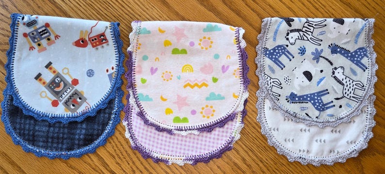 May include: Three baby bibs with different patterns. One has robots, another has stars and rainbows, and the third has zebras and giraffes. Each bib has a patterned bottom and is trimmed with lace.