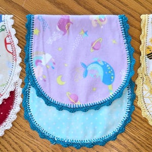 May include: Three baby bibs with scalloped edges. One bib features a red base with white polka dots and a top layer with vehicles. Another has a purple base with a blue polka dot layer and a space-themed top. The third has a yellow base with a chevron pattern and a bee-themed top.