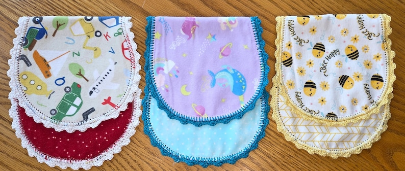 May include: Three baby bibs with scalloped edges. One bib features a red base with white polka dots and a top layer with vehicles. Another has a purple base with a blue polka dot layer and a space-themed top. The third has a yellow base with a chevron pattern and a bee-themed top.