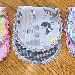 May include: Three baby bibs with different patterns and colors. One bib has a patchwork design with elephants, giraffes, and butterflies. Another has a gray and white woodland animal print. The third features a floral pattern. Each bib has a pink crocheted trim.