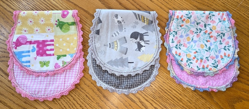 May include: Three baby bibs with different patterns and colors. One bib has a patchwork design with elephants, giraffes, and butterflies. Another has a gray and white woodland animal print. The third features a floral pattern. Each bib has a pink crocheted trim.