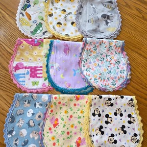May include: A collection of baby bibs with various patterns, including bees, animals, and floral designs. Each bib features a scalloped crochet trim in colors like yellow, pink, and blue. The bibs are made of soft fabric and are designed for infants.