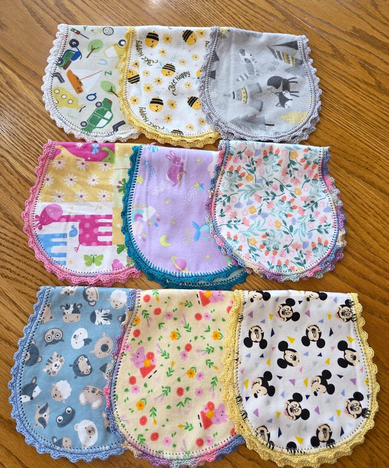 May include: A collection of baby bibs with various patterns, including bees, animals, and floral designs. Each bib features a scalloped crochet trim in colors like yellow, pink, and blue. The bibs are made of soft fabric and are designed for infants.