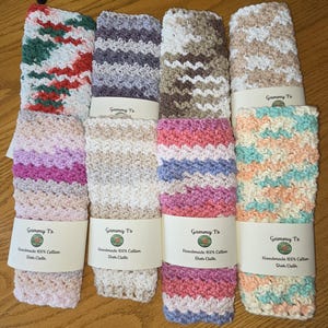 May include: A collection of handmade 100% cotton dish cloths in various colors and patterns. The cloths are folded and wrapped with labels that read "Gammy T's" and "Handmade 100% Cotton Dish Cloth". The patterns include stripes, zigzags, and color gradients.