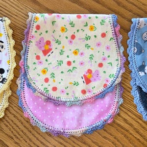 May include: Three baby bibs with decorative crochet edges. One bib features a Mickey Mouse pattern, another has a floral and butterfly design, and the third displays an animal print. Each bib has a different colored backing.