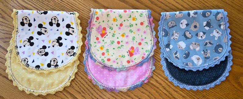 May include: Three baby bibs with decorative crochet edges. One bib features a Mickey Mouse pattern, another has a floral and butterfly design, and the third displays an animal print. Each bib has a different colored backing.