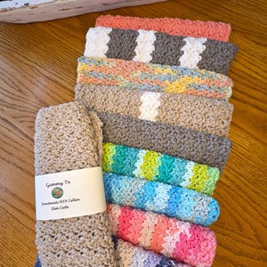 May include: A stack of handmade 100% cotton dish cloths in various colors, including pink, blue, green, and gray. One cloth is folded and has a label that reads "Gammy's Handmade 100% Cotton Dish Cloth."