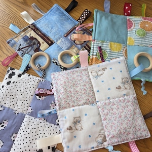 May include: A collection of three square fabric sensory blankets featuring patchwork designs with various patterns including kittens, floral prints, citrus slices, and cow spots, each adorned with multiple colorful ribbon loops and wooden rings.