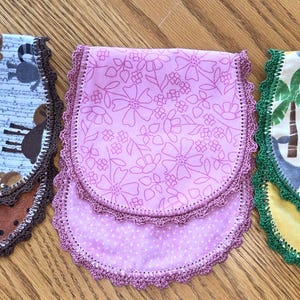 May include: Three handmade fabric bibs with scalloped edges. One features woodland animals, another has a floral pattern, and the third showcases a crocodile and elephant. Each bib is approximately 8 inches wide and 10 inches long.