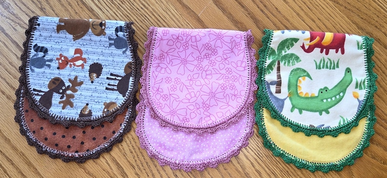 May include: Three handmade fabric bibs with scalloped edges. One features woodland animals, another has a floral pattern, and the third showcases a crocodile and elephant. Each bib is approximately 8 inches wide and 10 inches long.