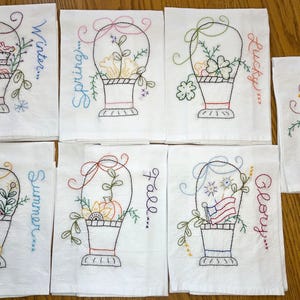 May include: A collection of white embroidered kitchen towels. Each towel features a basket design with seasonal themes like winter, spring, summer, and fall, along with the words "Lucky" and "Glory". The embroidery is in various colors.