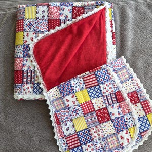 May include: A patchwork quilt and matching burp cloth. The quilt features a red, white, blue, and yellow patchwork design with a red interior and white lace trim. The burp cloth has a similar patchwork design.