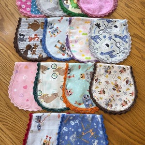 May include: A collection of baby bibs with various patterns and colors. Designs include animals, robots, and floral prints. Each bib has a contrasting crocheted edge in colors like pink, green, and brown. The bibs are arranged on a wooden surface.