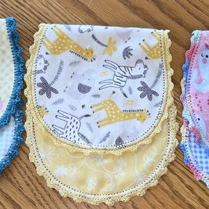May include: Three handmade baby bibs with colorful patterns and crochet edging. One bib features a yellow gingham pattern with cartoon vehicles, another has a white background with giraffes and zebras, and the third has a pink background with whales.
