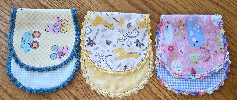 May include: Three handmade baby bibs with colorful patterns and crochet edging. One bib features a yellow gingham pattern with cartoon vehicles, another has a white background with giraffes and zebras, and the third has a pink background with whales.