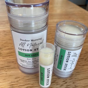 Puede incluir: Tres envases de barra de loción de plástico transparente con etiquetas blancas y detalles verdes. Las etiquetas dicen "Dusker Morning All Natural LOTION STICK BUG AWAY". El envase más grande está etiquetado "Net 57g."