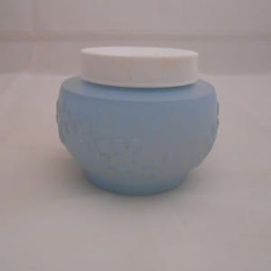 May include: A light blue, round jar with a white lid. The jar has a textured floral pattern around the body. The lid is white and has a ribbed texture. The jar is sitting on a white surface.