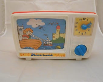 Pode incluir: Um relógio de brinquedo de plástico branco em forma de televisão vintage. O ecrã apresenta uma cena de desenho animado colorida com um barco, um castelo e nuvens. O texto "Musical toyclock" está impresso abaixo do ecrã. Mostrador amarelo.