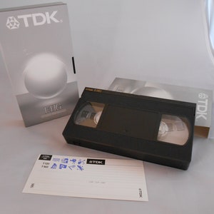 May include: A black VHS video cassette tape with a TDK label. The tape is in its original packaging, which is a silver and gray box with the TDK logo and the text "EHG Extra High Grade".