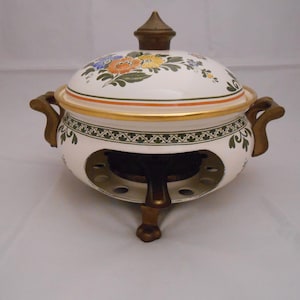 May include: A white ceramic tureen with a matching lid, featuring hand-painted orange, yellow, and blue floral motifs. The vessel has gold-toned trim, two side handles, and rests on a dark bronze-colored metal stand with three feet.