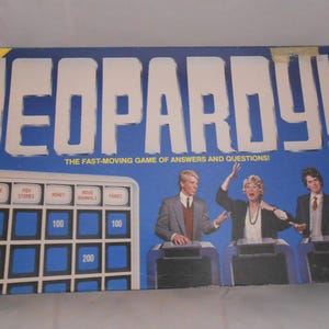 May include: Vintage Jeopardy! board game box with the game title in large white letters on a blue background. The box features a game board graphic and images of three people. The text "THE FAST-MOVING GAME OF ANSWERS AND QUESTIONS!" is also visible.