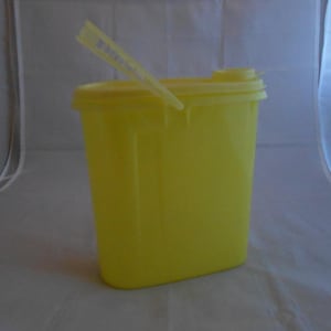 May include: A bright yellow plastic container with a lid and a small, attached spout. The container is rectangular with rounded corners and a handle-like feature on the lid. A yellow plastic stick is inserted into the lid.