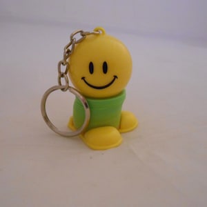 May include: A yellow smiley face keychain with a green body and yellow feet. The keychain has a silver chain and ring. The smiley face has black eyes and a curved smile. The keychain is made of plastic.