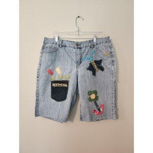 May include: Light blue denim shorts with embroidered floral and geometric designs. The shorts have a frayed hem and are decorated with colorful fabric appliques.