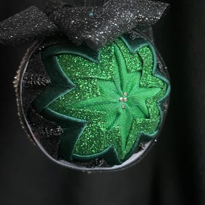 Peut inclure: Une boule de Noël verte et noire avec un motif en forme d'étoile et des paillettes. La boule est décorée d'un ruban noir.