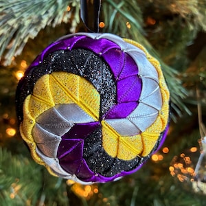 Op de afbeelding: Een handgemaakte kerstbal met een zwart lint om op te hangen. De bolvormige versiering heeft secties van paarse, gele, zilveren en zwarte glitterstof. Hij hangt voor een vage kerstboom.