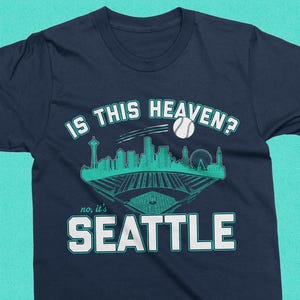 May include: Navy blue t-shirt with a teal graphic of the Seattle skyline, a baseball, and the text "IS THIS HEAVEN?" and "no, it's SEATTLE". The design includes the Space Needle and a baseball field.