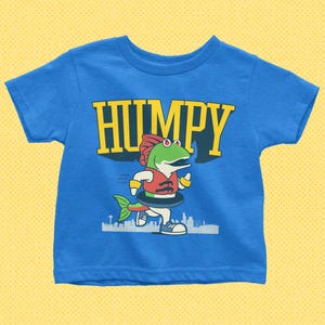 May include: A blue t-shirt featuring the word "HUMPY" in yellow block letters above a cartoon fish character. The fish is wearing a red vest and a swim ring, with a cityscape silhouette at the bottom.