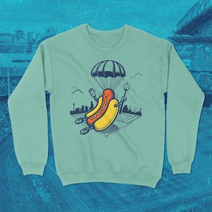 Puede incluir: Sudadera de color verde azulado claro con un personaje de dibujos animados de perrito caliente con paracaídas y tridente, flotando sobre un paisaje urbano estilizado y un campo de béisbol. El diseño es juguetón y caprichoso.