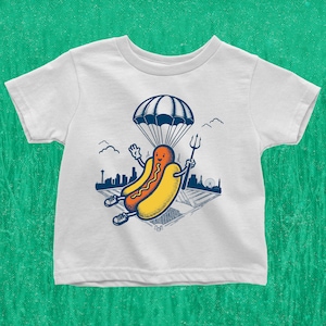 Puede incluir: Camiseta blanca con un gráfico de un perrito caliente con un paracaídas y sosteniendo una lanza. El perrito caliente está sonriendo y tiene una ciudad en el fondo.