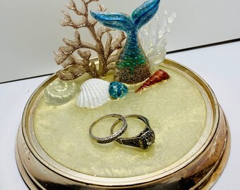 Puede incluir: Un porta anillos de oro con una cola de sirena, coral, conchas marinas y dos anillos sobre una base de arena blanca.