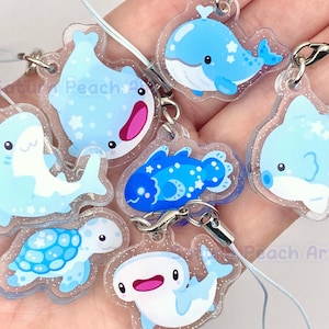 May include: A collection of clear acrylic charms featuring various marine animals in shades of blue, including a whale, shark, and turtle. Each charm has a glittery border and a silver clasp for attachment. The charms are designed with cartoon-like illustrations.