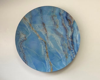 Pode incluir: Uma decoração de parede redonda com um padrão de mármore azul e dourado. A decoração está pendurada em uma parede branca acima de uma mesa de mármore preto com três livros empilhados um em cima do outro.