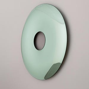 May include: A modern, oval-shaped wall mirror with a central circular cutout. The mirror has a soft, mint-green hue and a black border. The design is minimalist and contemporary, suitable for various interior styles.