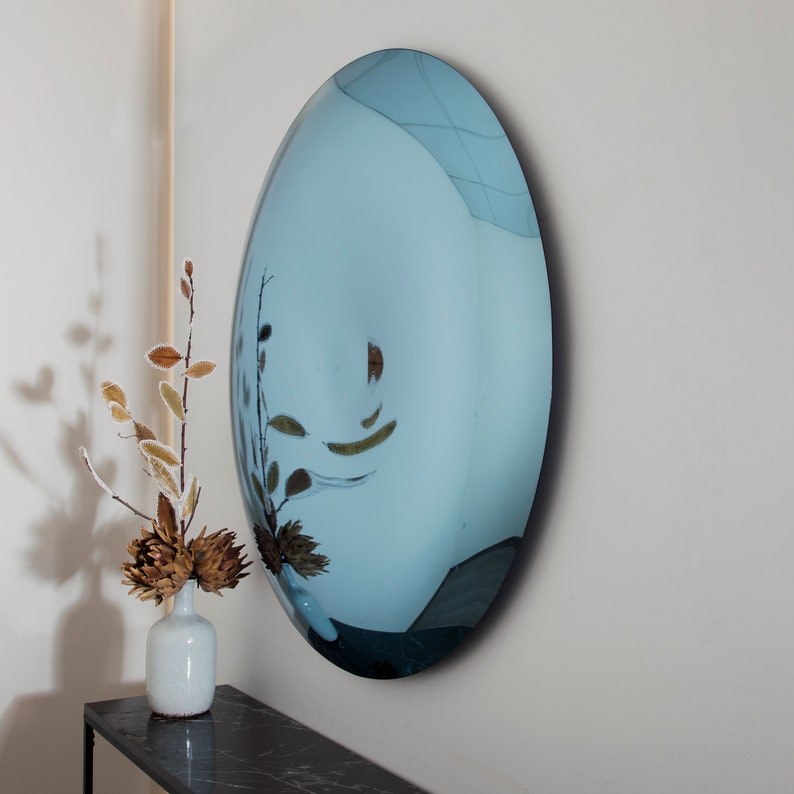Puede incluir: Un espejo grande, redondo y te&ntilde;ido de azul cuelga de una pared blanca. El espejo es ligeramente convexo, creando un reflejo distorsionado de la habitaci&oacute;n. Un jarr&oacute;n con ramas secas se encuentra sobre una mesa negra frente al espejo.
