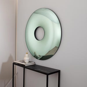 May include: A large, round mirror with a central hole, mounted on a light gray wall. The mirror has a green-tinted surface and a black frame. A black table with a marble top and a decorative jar are below the mirror.
