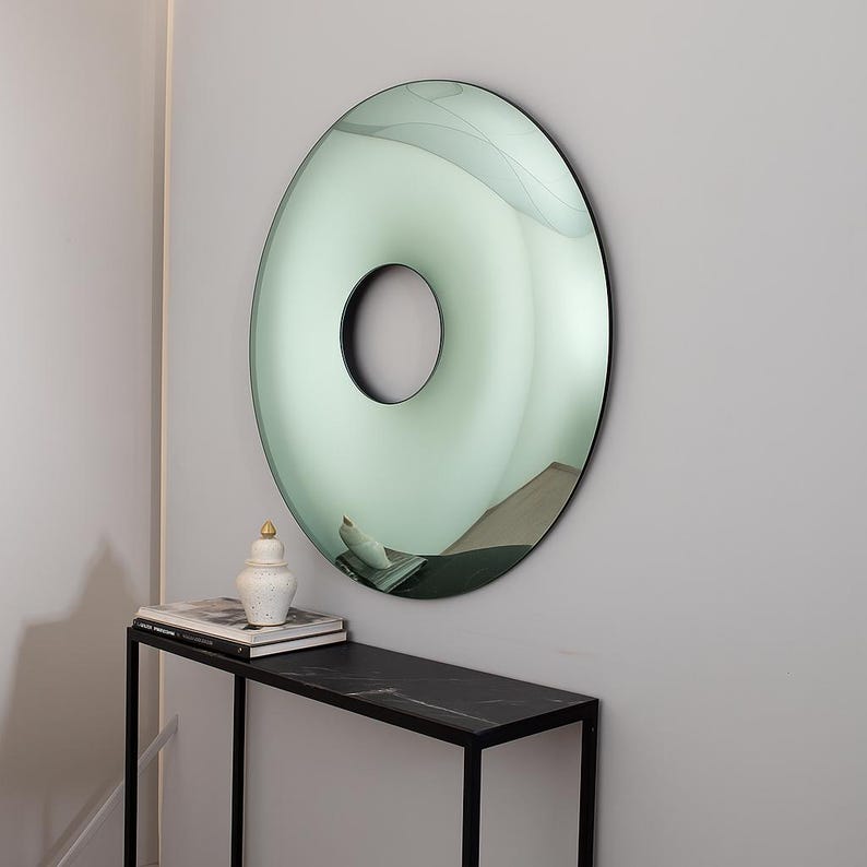 May include: A large, round mirror with a central hole, mounted on a light gray wall. The mirror has a green-tinted surface and a black frame. A black table with a marble top and a decorative jar are below the mirror.