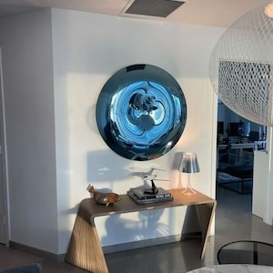 Puede incluir: Un espejo de pared redondo azul reflectante con una superficie ondulada cuelga de una pared blanca sobre una mesa consola de madera. Un peque&ntilde;o avi&oacute;n modelo, un libro titulado "Royal Oak" y una figurita de pescado decorativa est&aacute;n sobre la mesa. Una l&aacute;mpara de mesa plateada se encuentra sobre la mesa.