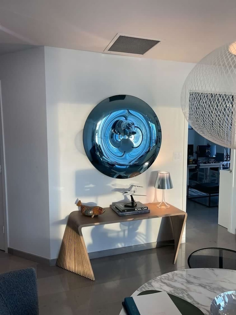 Puede incluir: Un espejo de pared redondo azul reflectante con una superficie ondulada cuelga de una pared blanca sobre una mesa consola de madera. Un peque&ntilde;o avi&oacute;n modelo, un libro titulado "Royal Oak" y una figurita de pescado decorativa est&aacute;n sobre la mesa. Una l&aacute;mpara de mesa plateada se encuentra sobre la mesa.