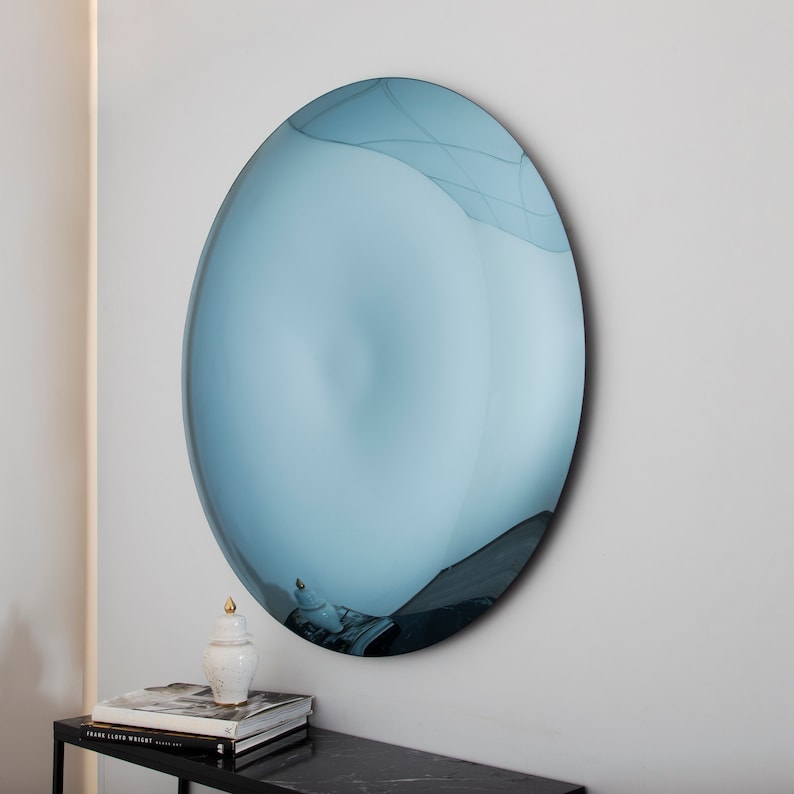 Puede incluir: Un espejo grande, redondo, de color azul con una superficie ligeramente convexa. El espejo est&aacute; colgado en una pared blanca.