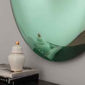 May include: A decorative arrangement featuring a large, round, mint-green mirror with a gold-topped ceramic jar and a smaller matching jar. The jars sit on a black table with books.