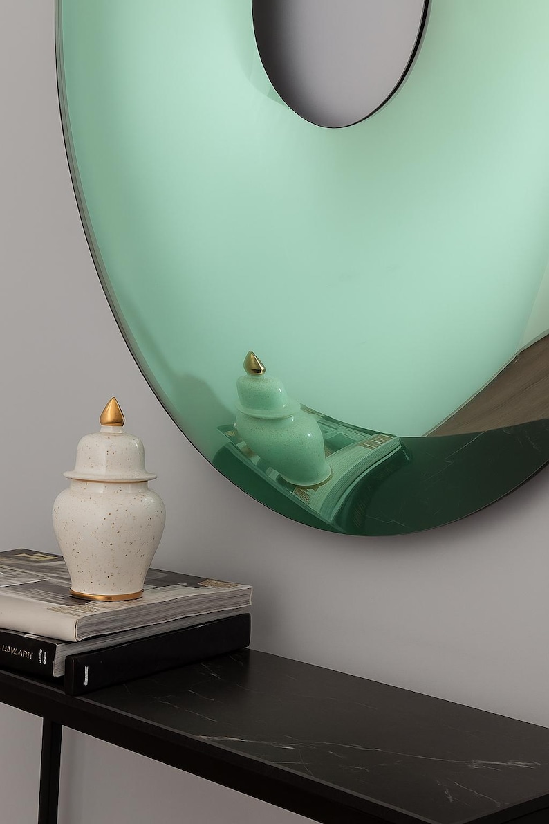 May include: A decorative arrangement featuring a large, round, mint-green mirror with a gold-topped ceramic jar and a smaller matching jar. The jars sit on a black table with books.