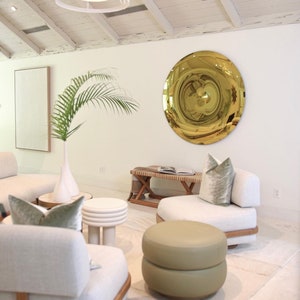 Pode incluir: Uma sala de estar com paredes brancas e um teto de madeira branca. H&aacute; um grande espelho redondo dourado na parede. H&aacute; dois sof&aacute;s brancos com almofadas verdes e um pufe verde no centro da sala.