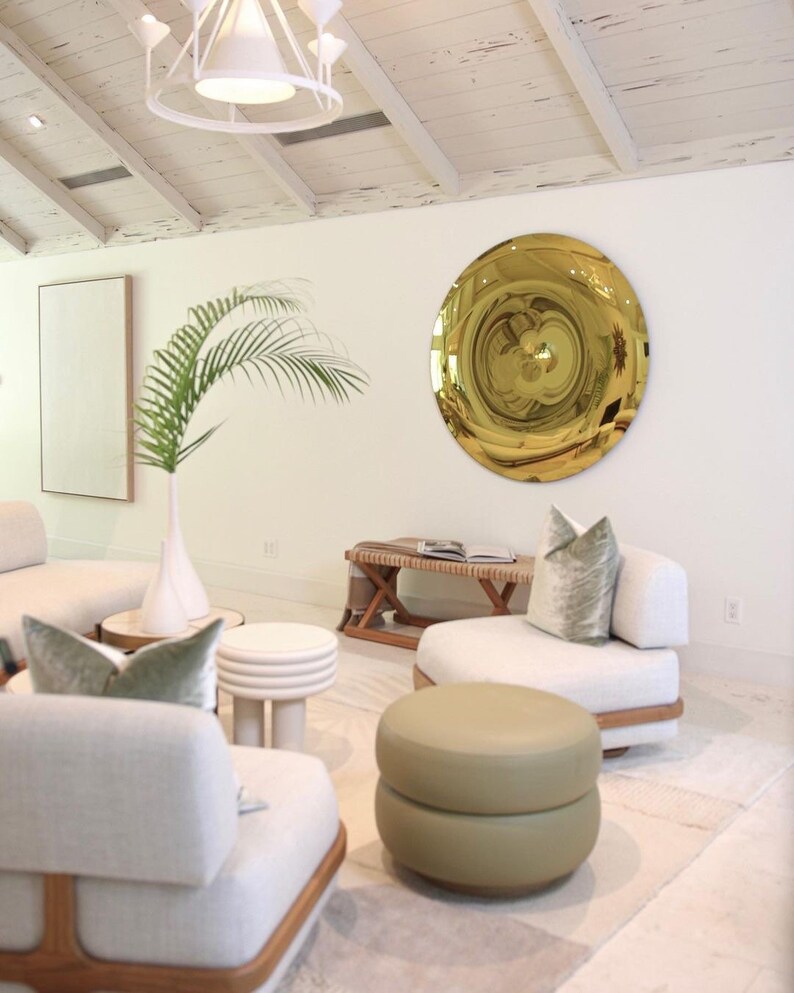 Pode incluir: Uma sala de estar com paredes brancas e um teto de madeira branca. H&aacute; um grande espelho redondo dourado na parede. H&aacute; dois sof&aacute;s brancos com almofadas verdes e um pufe verde no centro da sala.